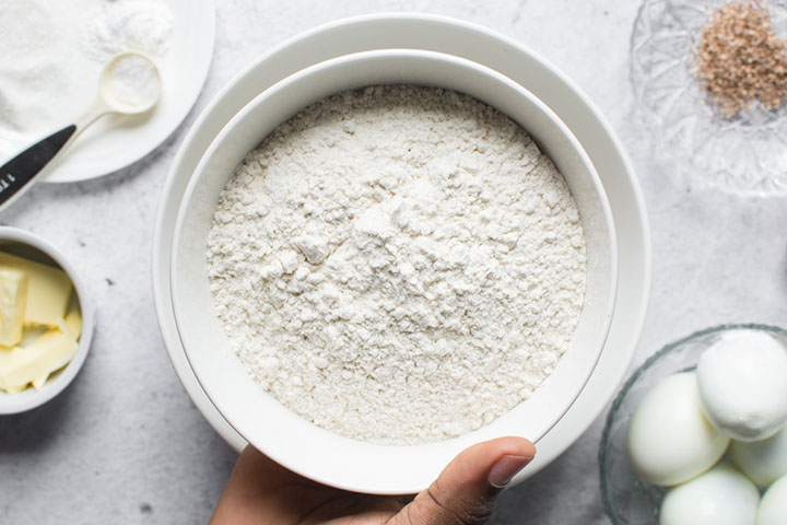 hand holding a white bowl containing flour surrounded by saucers and bowls containing ingredients
